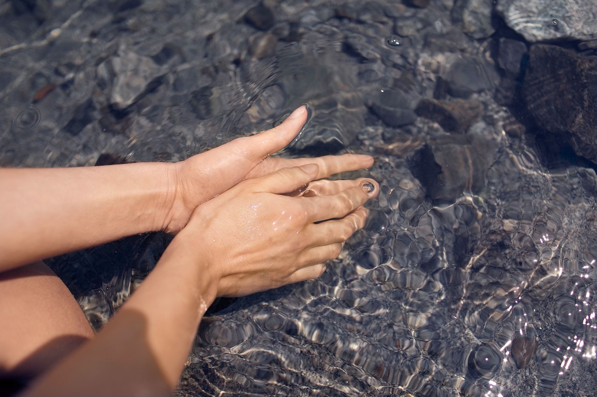Hände in klarem Wasser. Schwarze Fingernägel. Wasser mit Steinen am Boden.